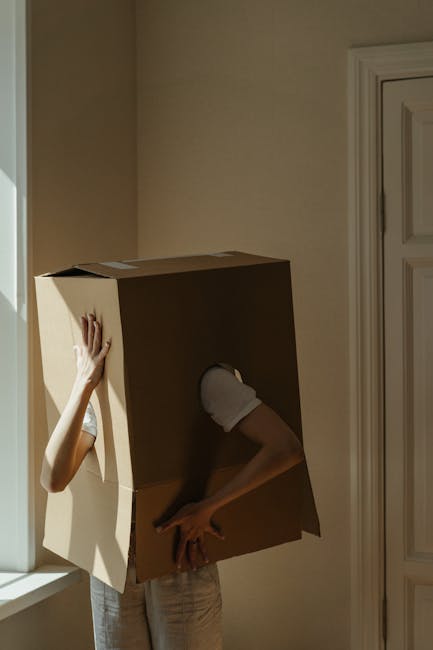 A smiling man with a curly afro hairstyle and a beard stands inside a bright room with large arched windows, holding a medium-sized cardboard moving box in his right arm while making an 'OK' gesture w