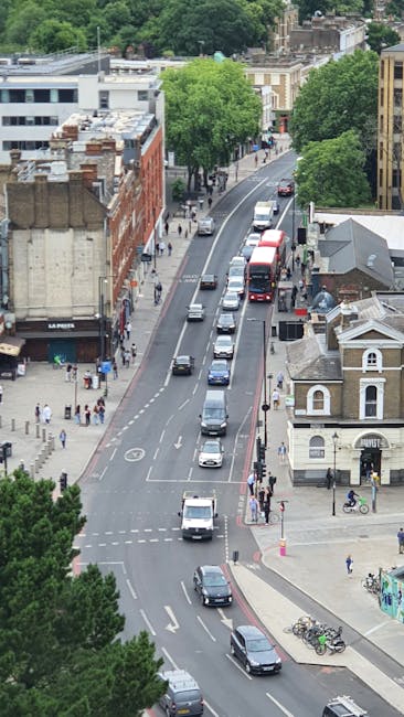 An aerial view of a narrow urban street in Catford, showing a line of parked cars along the curb and moving vehicles on the road, with pedestrians walking on the sidewalks and crossing at designated crosswalks. On the left side, there are multi-storey red brick buildings, some with storefronts, while on the right side, there is a small white building with a gabled roof and a bicycle rack nearby. Lush green trees line both sides of the street, providing shade and greenery in the area. The scene captures a typical busy street during daytime, highlighting the challenges of moving furniture and household items through a narrow access route, which Catford Movers may expertly handle as part of their home relocation and furniture transport services. The image underscores the importance of careful planning for removals in constrained city environments, with the street activity and urban infrastructure visible.