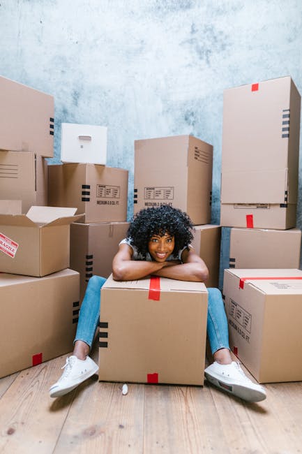 A young woman with curly black hair is sitting on a wooden floor in a room filled with numerous cardboard moving boxes of various sizes, some sealed with red tape and others partially opened. The boxes are stacked against a textured, light blue wall, creating a cluttered environment associated with home relocation. The woman is smiling, resting her chin on her crossed arms placed on a medium-sized box, and wearing casual clothing including a light-colored top and jeans with white sneakers. This scene depicts packing and moving activities typical of furniture transport and house removals, with the boxes arranged systematically indicating an organized packing process by Catford Movers for a relocation near Mountsfield Park. Visible in the room are the cardboard boxes, some with labels, and the natural lighting highlights the orderly yet busy atmosphere of preparing for a house move.