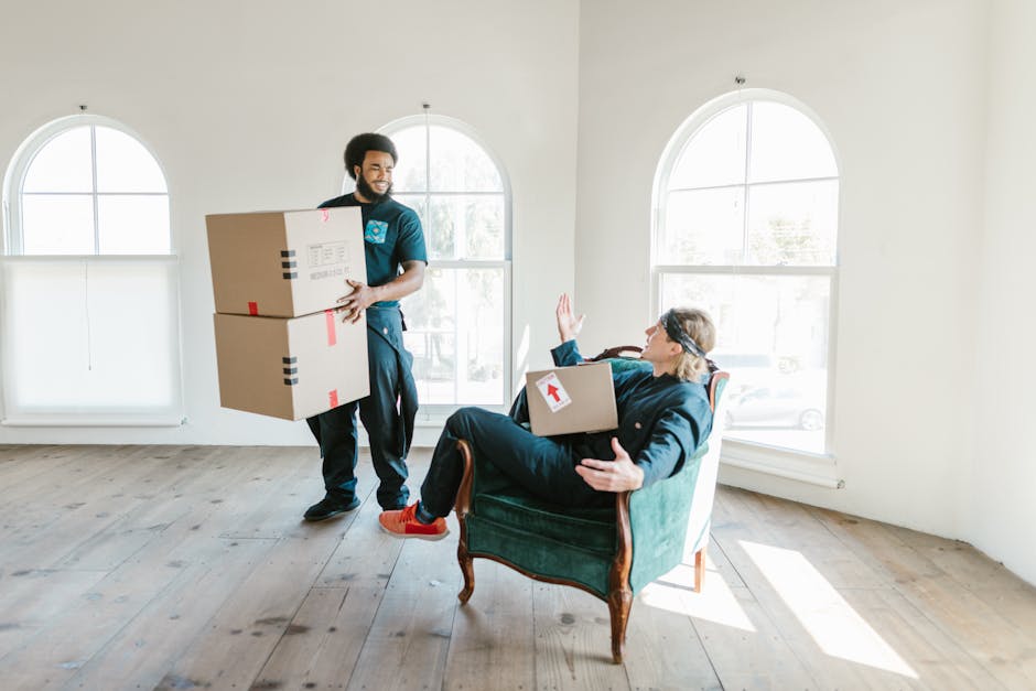 A professional mover from Catford Movers is inside a bright, spacious room with light-colored walls and large, arched windows, engaged in packing and home relocation activities. The mover, dressed in uniform, is holding two cardboard boxes with red tape, preparing to move them. A woman, seated in an elegant green upholstered armchair, is holding a smaller box on her lap, with her left hand raised as if signaling to stop or communicate, while wearing casual clothing and red sneakers. The room features wooden flooring and ample natural daylight streaming through the windows, creating a well-lit environment suitable for furniture transport and packing during a house removal. The scene captures the ongoing loading process involved in furniture transport or moving services, emphasizing careful handling and the organization involved in house removals by Catford Movers.