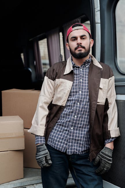 A young man with a beard and mustache, wearing a red cap backward, a beige and brown jacket over a blue checkered shirt, and grey gloves, standing inside the back of a moving van. Behind him are several cardboard boxes of various sizes, some stacked and others placed side by side, all ready for a home relocation. The van's interior is lined with dark fabric, and the environment is well-lit, indicating daytime. The scene depicts the process of furniture transport and packing during a house move, with the man preparing boxes for loading or unloading, illustrating essential steps in packing and moving services provided by Catford Movers as part of a smooth Catford move process. The focus is on the loading process, showing the careful handling of household goods in preparation for transportation.