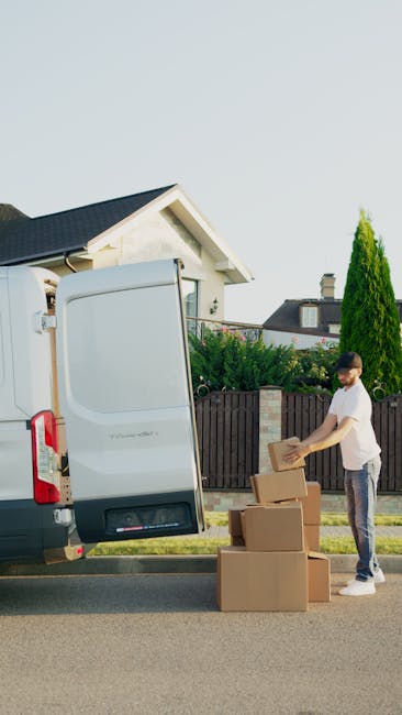 A man and a woman stand beside an open vehicle, such as a van or moving truck, loaded with numerous cardboard boxes of various sizes, some stacked on top of each other. The man, dressed in a white t-shirt, dark cap, and wearing wireless earbuds, is holding a clipboard and pen, possibly taking inventory or notes related to the moving process. The woman, wearing a white cap, black top, and light blue jeans, holds one of the larger cardboard boxes, preparing for furniture transport or packing during a home relocation. The scene takes place outdoors, near a paved area, with natural lighting illuminating the interior of the vehicle, which is filled with securely packed boxes and some packing materials like plastic wrap. The environment and actions suggest a professional moving process, with attention to careful packing and effective loading, consistent with a house removals or relocation service such as those offered by Catford Movers, supporting the topic of smooth Catford moves and packing tips.