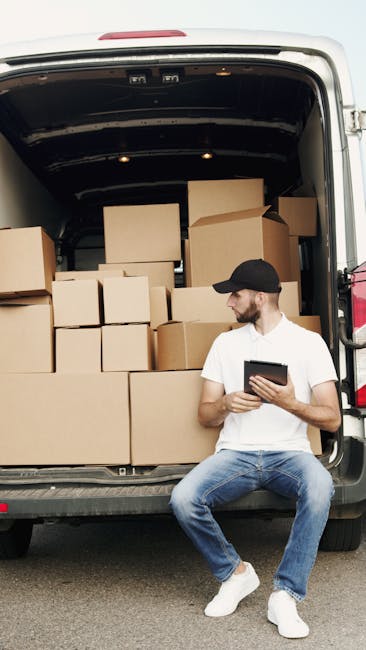 A man wearing a white t-shirt, jeans, and a black cap sits on the edge of a loaded moving van with its rear door open. Inside the van, numerous cardboard boxes of various sizes are arranged, some stac