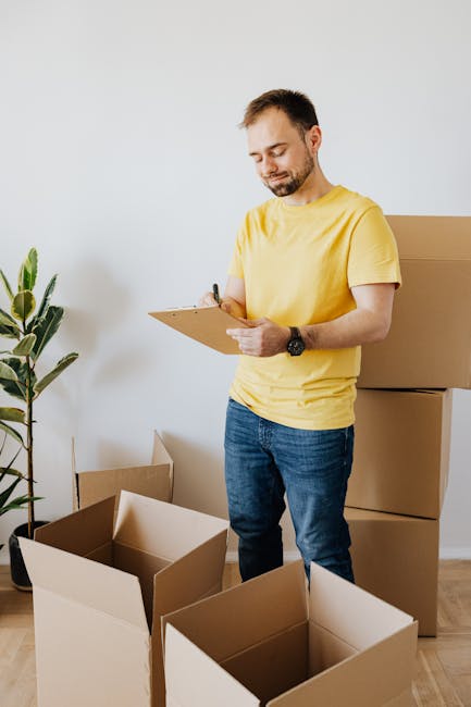 A professional removalist from Catford Movers, dressed in a dark blue t-shirt, is inside a room filled with moving boxes made of cardboard and plastic wrapping, reviewing a checklist on a clipboard. T